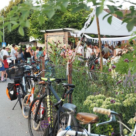 Viele Fahrräder sind in der Nähe eines Gartens mit einem weißen Baldachin geparkt, wo mehrere Menschen plaudern. Bäume und Pflanzen sind im Hintergrund.