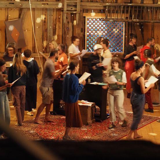 Eine Gruppe von Menschen versammelt sich in einem Raum mit Holzwänden. Sie halten Papiere in den Händen und scheinen ein Theaterstück zu proben. Ein Teppich bedeckt den Boden und ein Klavier steht in der Nähe. Ein Fenster mit einem rautenförmigen Gitter befindet sich im Hintergrund.