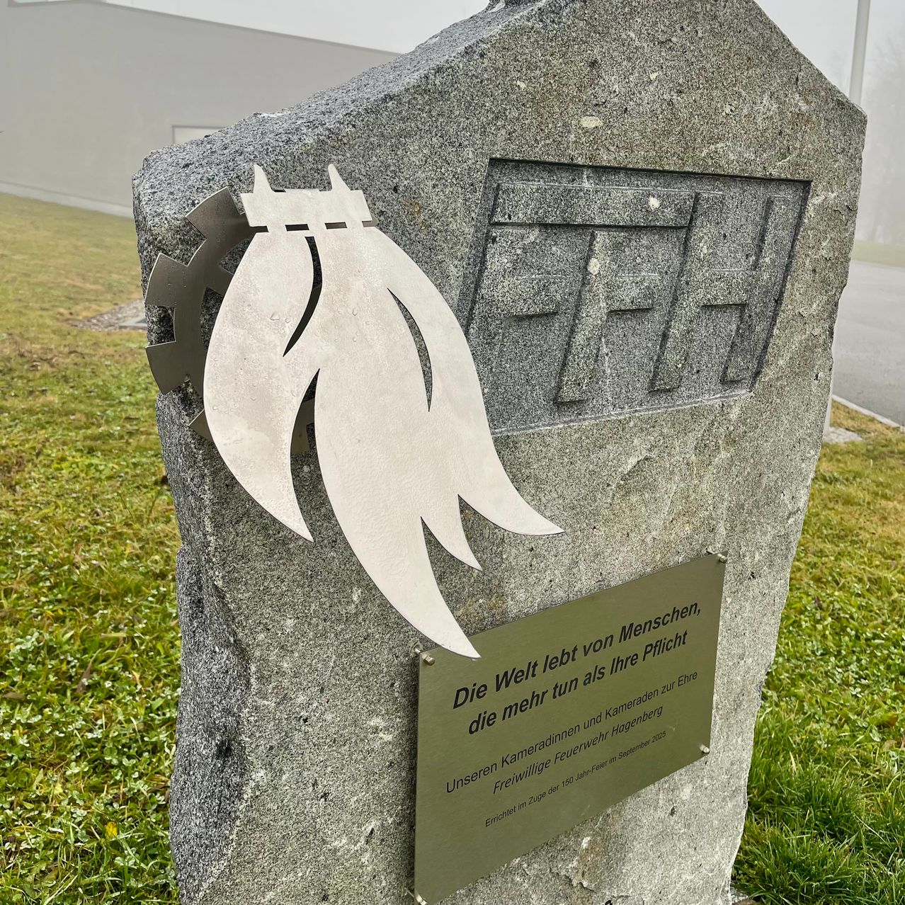 Ein Steinmonument mit einem weißen Metalldesign und Text auf Deutsch. Es steht 'Die Welt lebt mehr als deine Pflicht als dein Recht.' Eine Plakette erinnert an die Freiwillige Feuerwehr Hamburg.