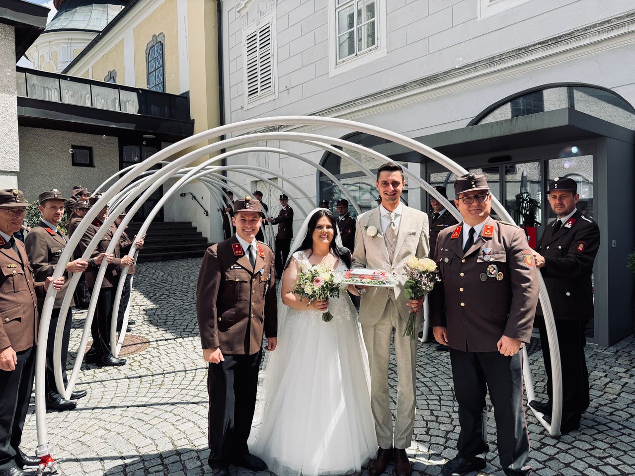 Ein frisch verheiratetes Paar und zwei Militäroffiziere stehen lächelnd unter einem Torbogen vor einem Gebäude. Der Bräutigam trägt einen hellen Anzug, und die Braut hält Blumen. Dahinter stehen mehrere Offiziere als Wache.