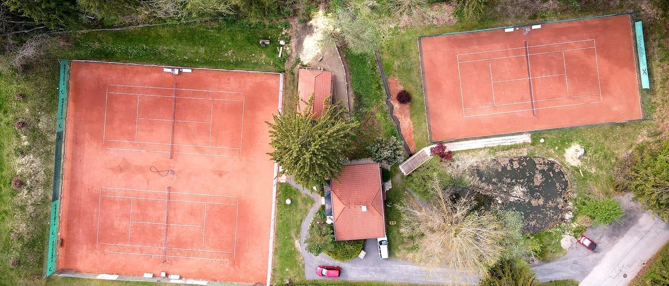 Ein Luftbild eines Tennisplatzkomplexes zeigt zwei Tennisplätze mit roten Tonoberflächen. Die Plätze sind von üppigem Grün umgeben. Ein kleines Haus mit rotem Dach befindet sich zwischen den Plätzen, und ein Teich ist in der Nähe. Ein rotes Auto ist auf dem Einfahrtsweg geparkt.