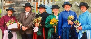 Fünf Menschen in traditioneller Kleidung stehen draußen. Sie halten Blumen und lächeln. Dahinter befindet sich eine Steinmauer und ein Holzzaun.