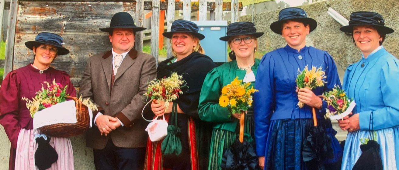 Fünf Menschen in traditioneller Kleidung stehen draußen. Sie halten Blumen und lächeln. Dahinter befindet sich eine Steinmauer und ein Holzzaun.