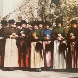 Eine Gruppe von Frauen in traditioneller Kleidung, die Blumen und Regenschirme halten, steht vor einem Gebäude mit Bäumen im Hintergrund.