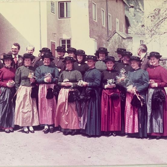 Eine Gruppe von Frauen und Männern in traditioneller Kleidung posiert für ein Foto vor einem Gebäude. Die Frauen tragen Hüte und halten Blumensträuße, während die Männer dahinter in Anzügen und Krawatten stehen.
