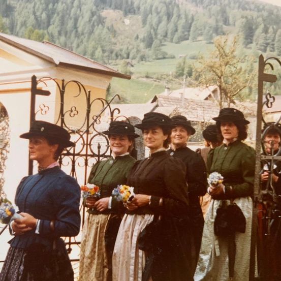 Eine Gruppe von Frauen in traditioneller Kleidung steht lächelnd und Blumen haltend draußen. Sie stehen vor einem Tor, mit einem Berg und Bäumen im Hintergrund.