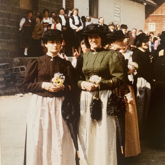 Eine Gruppe von Frauen in traditioneller Kleidung steht vor einem Gebäude mit einer Menschenmenge im Hintergrund.