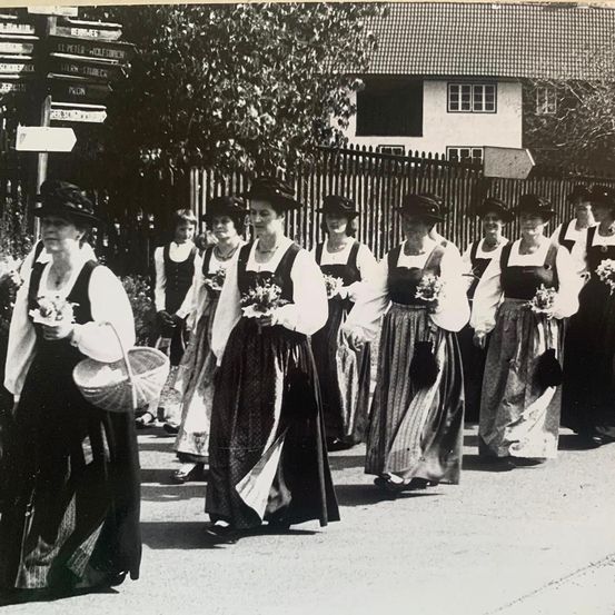 Eine Gruppe von Frauen in traditioneller Kleidung, wahrscheinlich an einem Umzug teilnehmend, geht in einer Reihe. Sie halten Körbe und Blumen.