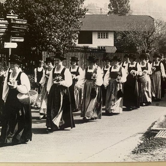 Schwarz-Weiß-Bild einer Parade mit Frauen in traditionellen Kostümen, die Blumen und Körbe tragen, die vor einem Haus mit Zaun auf einer Straße laufen.