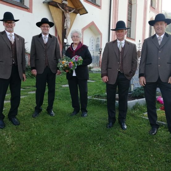 Vier Personen stehen auf dem Rasen vor einer Kirche. Drei Männer und eine Frau. Die Frau hält einen Blumenstrauß.