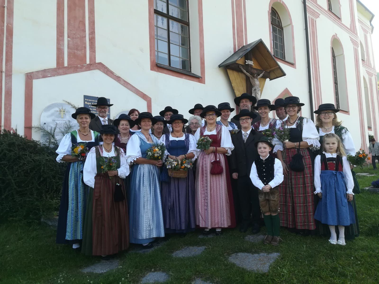 Eine Gruppe von Menschen in traditioneller bayerischer Kleidung, einschließlich Hüten und Kleidern, posiert für ein Foto vor einer Kirche mit einem Kruzifix über dem Eingang.