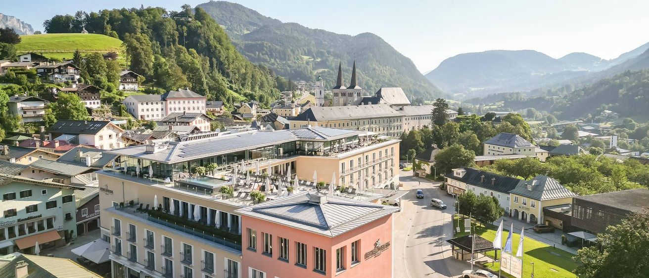 Luftaufnahme einer Stadt mit Bergen im Hintergrund, mit einem modernen Hotel, einer Kirche und einer Straße mit Autos und Fahnen.