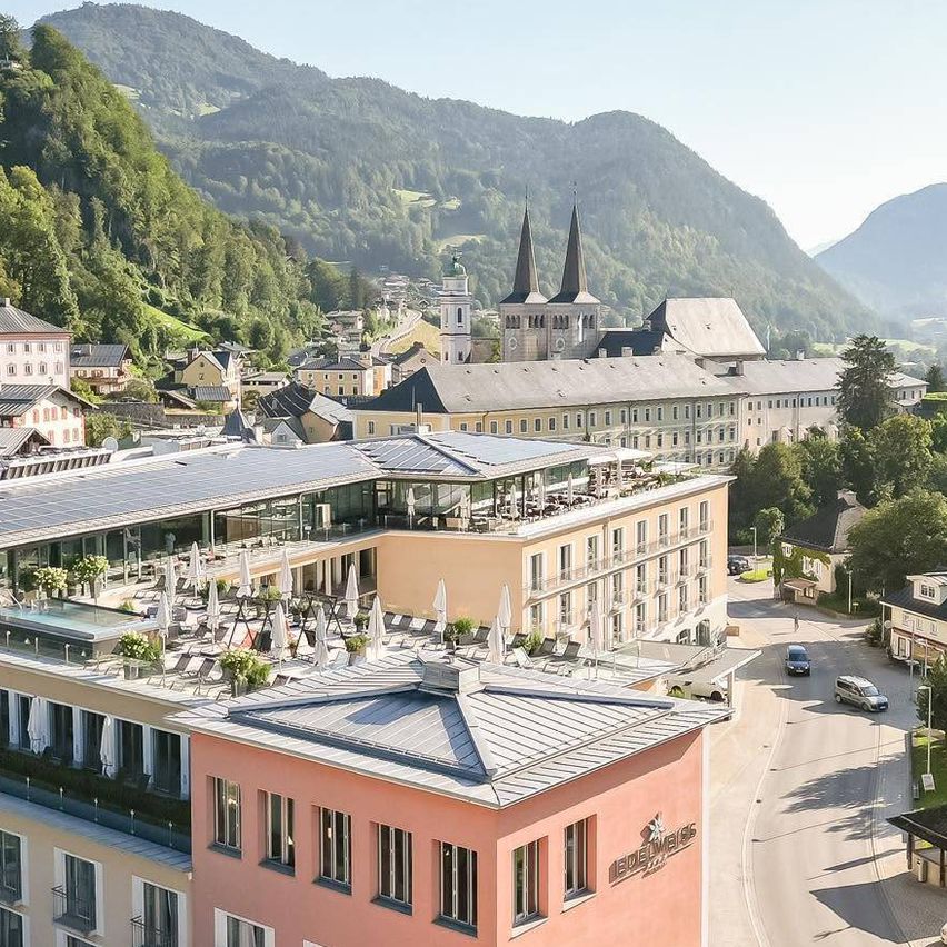 Luftaufnahme einer Stadt mit Bergen im Hintergrund, mit einem modernen Hotel, einer Kirche und einer Straße mit Autos und Fahnen.