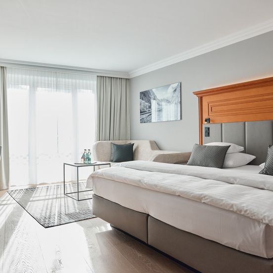 Ein modernes Schlafzimmer mit einem großen Bett, einem hölzernen Kopfteil und einer gemütlichen Couch in der Nähe eines Fensters. Der Raum hat einen Holzboden und weiße Wände.