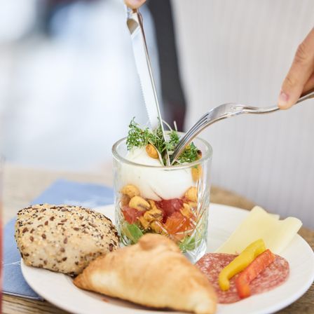 Eine Person benutzt Messer und Gabel, um ein Gericht mit Ei, Gemüse und Brot auf einem Teller zu essen.