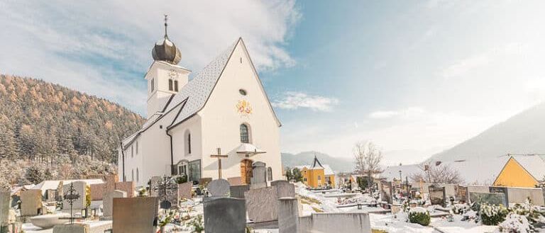 Eine Kirche mit goldener Kuppel steht in einem von Schnee bedeckten Friedhof, umgeben von Grabsteinen und einem gelben Gebäude im Hintergrund.