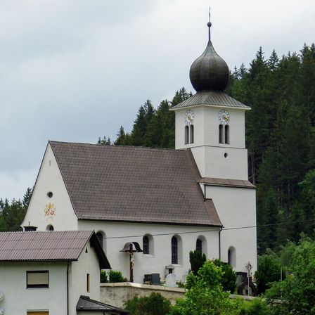 Eine weiße Kirche mit einem braunen Dach steht gegen einen bewölkten Himmel, umgeben von hohen Bäumen. Der Turm hat eine Uhr und eine Kuppel.
