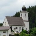 Eine weiße Kirche mit einem braunen Dach steht gegen einen bewölkten Himmel, umgeben von hohen Bäumen. Der Turm hat eine Uhr und eine Kuppel.