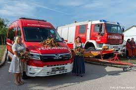 Zwei Frauen stehen neben einem neuen Feuerwehrauto an einem sonnigen Tag. Das Auto ist mit Sonnenblumen dekoriert. Ein weiteres Feuerwehrauto ist dahinter geparkt.
