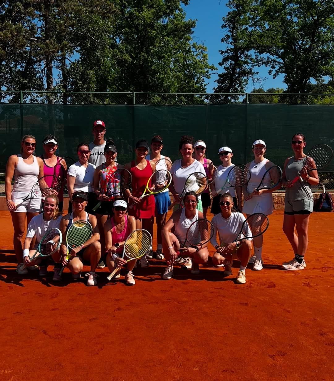 Eine Gruppe von Tennisspielern posiert für ein Foto auf einem Sandplatz mit Bäumen im Hintergrund.