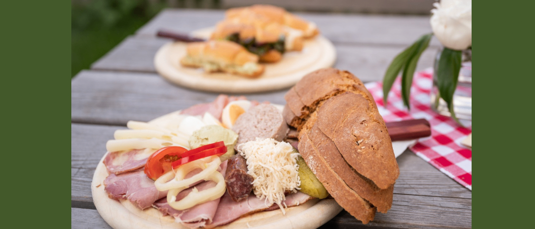 Ein grünes Banner mit 'Kleinschuster' Logo zeigt einen Holztisch mit zwei Essensplatten, eine mit einem Sandwich und eine andere mit einem Fleischplatten. Der Datumsbereich vom 31. Juli bis 31. August ist angegeben.