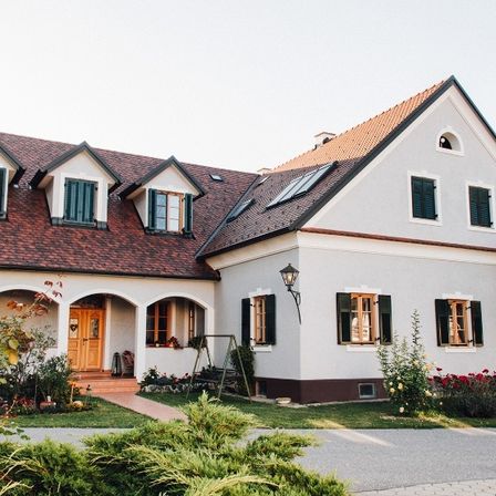 Ein weißes Haus mit einem braunen Dach und grünen Fensterläden, umgeben von einem Garten mit roten Blumen und einer Schaukel.