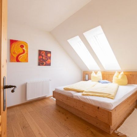 Ein gemütliches Schlafzimmer mit Holzboden, weißen Wänden und zwei dreieckigen Fenstern. Ein Bett mit gelben Kissen und Decken steht an der Wand. Zwei abstrakte Gemälde sind an der Wand montiert.