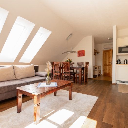 Ein gemütliches Dachgeschoss-Wohnzimmer mit Holzböden, einem Sofa, einem Couchtisch und einem Essbereich mit Tisch und Stühlen. Der Raum hat eine warme, einladende Atmosphäre mit natürlichem Licht, das durch die Fenster hereinkommt.