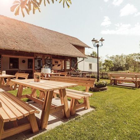 Ein rustikaler Essbereich im Freien mit Holztischen und -bänken unter einem strohgedeckten Dach, umgeben von saftigem grünem Gras und Bäumen.