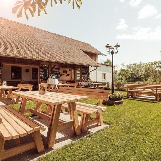 Ein rustikaler Essbereich im Freien mit Holztischen und -bänken unter einem strohgedeckten Dach, umgeben von saftigem grünem Gras und Bäumen.