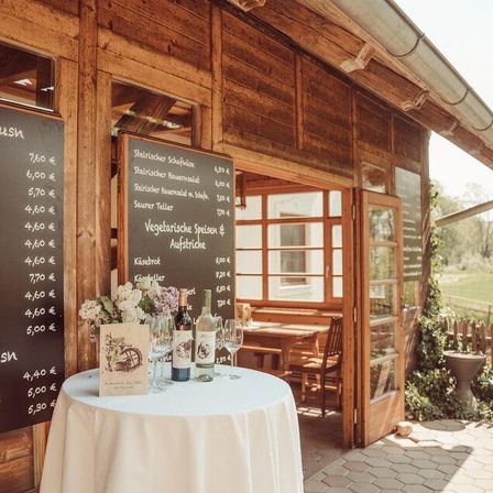 Ein rustikales Holzhaus mit einer Speisekarte, einem Tisch mit Weinflaschen und Blumen und einer offenen Tür, die zu einer Holzbank und einem Tisch mit Stühlen führt.