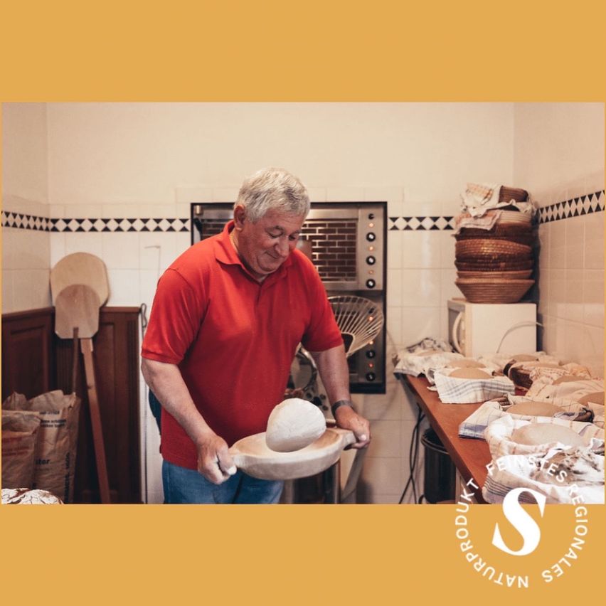 Ein Mann in einem roten Hemd arbeitet mit einem Steinmörser und Stößel in einer Küche. Verschiedene Schüsseln und Küchenutensilien sind im Hintergrund zu sehen.