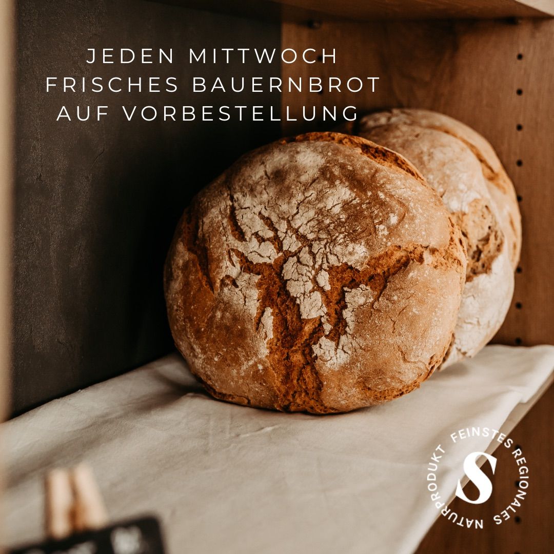 Ein Holzregal zeigt zwei Laibe rustikales, knuspriges Brot, mit einem weißen Tuch darunter und einem weißen Textüberlagert, das 'Jeden Mittwoch frisches Bauernbrot auf Vorbestellung' lautet.