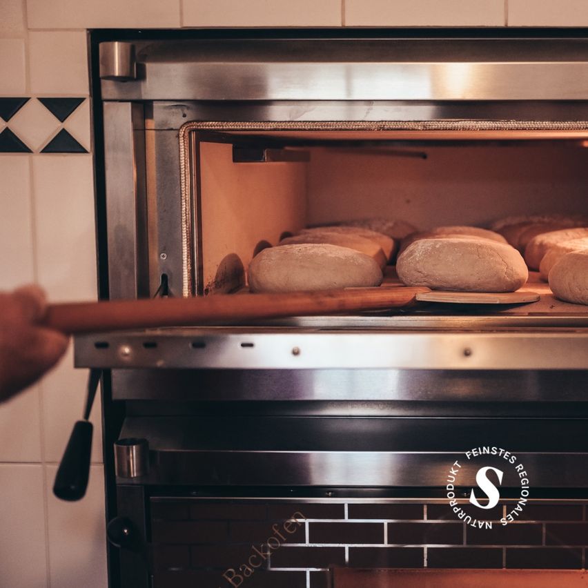 Eine Hand, die einen Holzspatel hält und ein Tablett mit Brot aus einem gewerblichen Ofen nimmt. Der Ofen hat eine geflieste Rückwand und ein Logo in der unteren rechten Ecke.