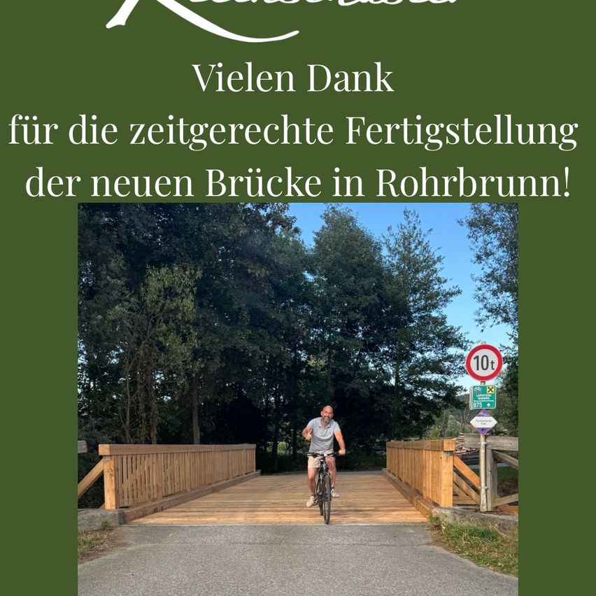 Ein Mann fährt auf einem hölzernen Steg, umgeben von Bäumen, mit einem Fahrrad. Ein Schild auf der rechten Seite zeigt ein Geschwindigkeitslimit von 10 km/h an. Der Hintergrundtext lautet 'Vielen Dank für die rechtzeitige Fertigstellung der neuen Brücke in Rohrbrunn!'.