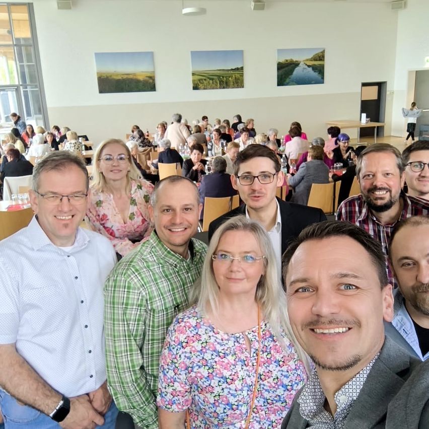 Eine Gruppe von Menschen posiert für ein Foto in einem großen Raum. Sie lächeln und schauen zur Kamera. Der Raum hat eine weiße Wand mit drei Gemälden.