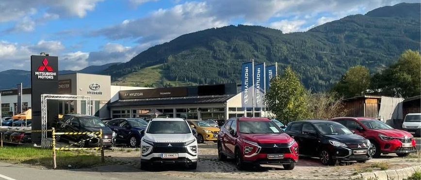 Ein Autohaus mit einem Mitsubishi Motors-Schild zeigt mehrere geparkte Autos davor. Hinter dem Autohaus sind Berge sichtbar.