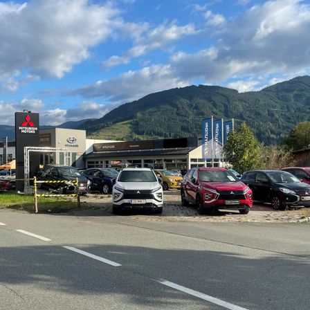 Ein Autohaus mit Mitsubishi- und Hyundai-Bannern. Vor dem Gebäude sind verschiedene Autos geparkt, darunter ein roter und ein weißer SUV. Im Hintergrund sind Berge zu sehen.