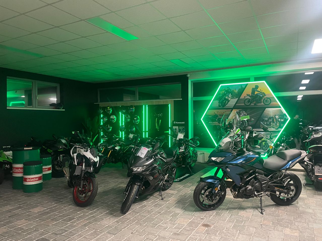 Ein Ausstellungsraum mit mehreren Motorrädern, die unter grünen Lichtern ausgestellt sind. Helme und Werbebilder schmücken die Wände.