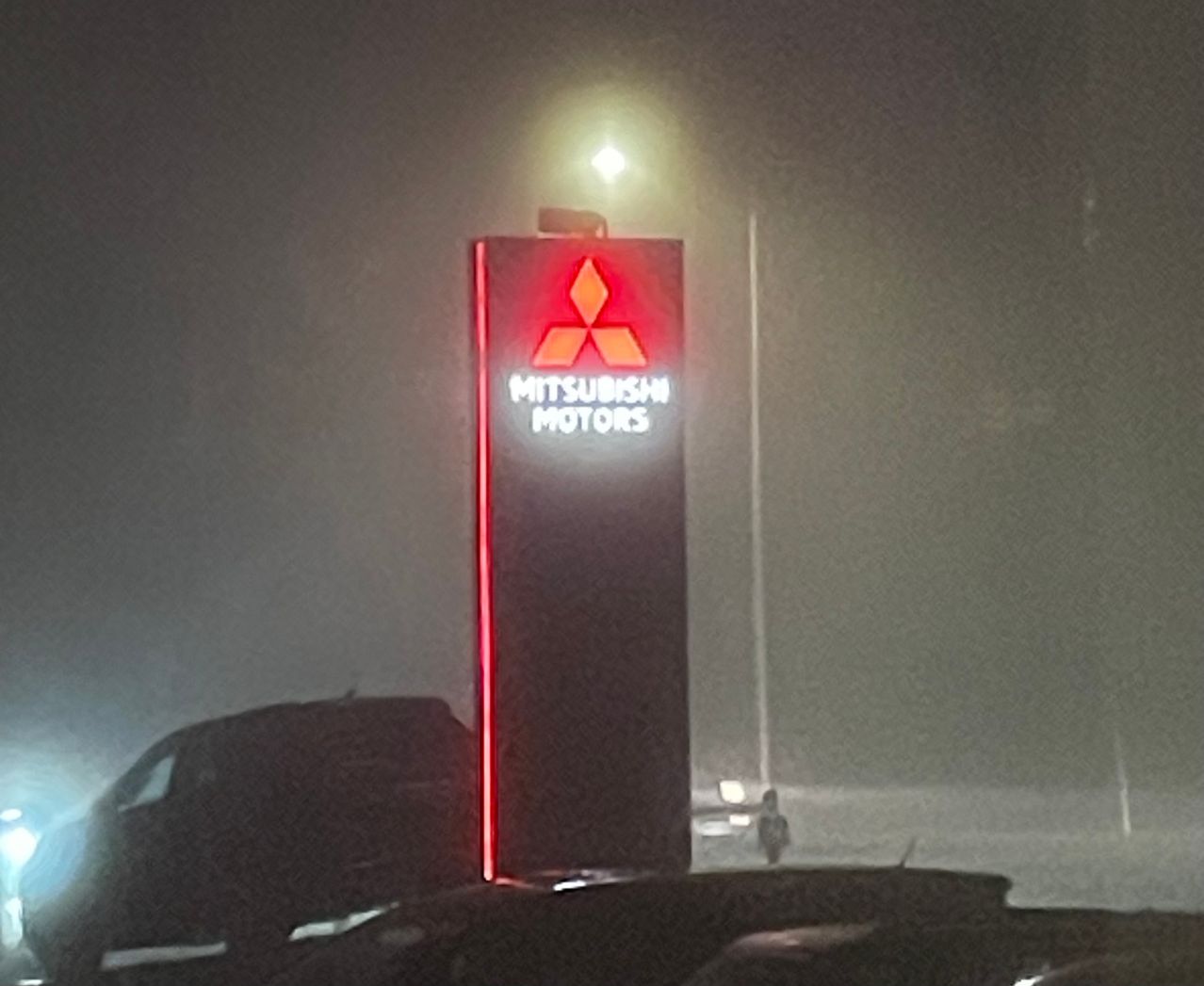 Ein Mitsubishi Motors-Schild ist im Nebel beleuchtet, ein Fahrzeug ist in der Nähe geparkt. Das Schild ist rot mit weißen Texten und steht aufrecht.