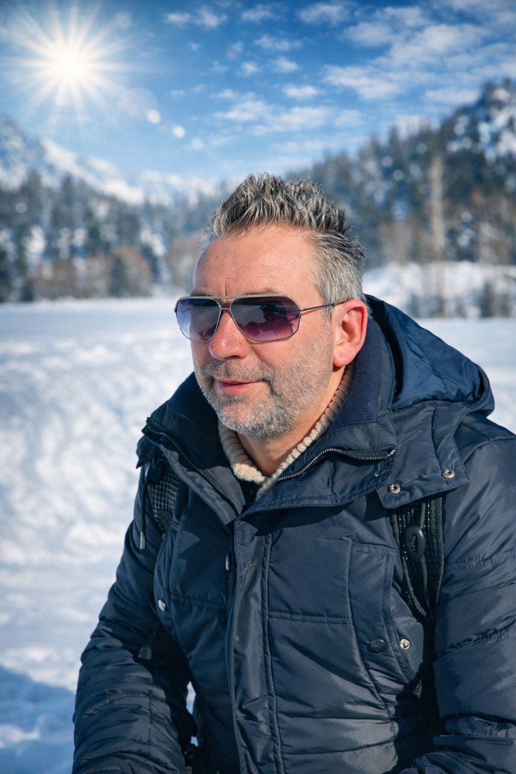 Ein Mann mit grauem Haar, der eine Sonnenbrille und eine blaue Jacke trägt, steht im Schnee mit einer Bergkette im Hintergrund.