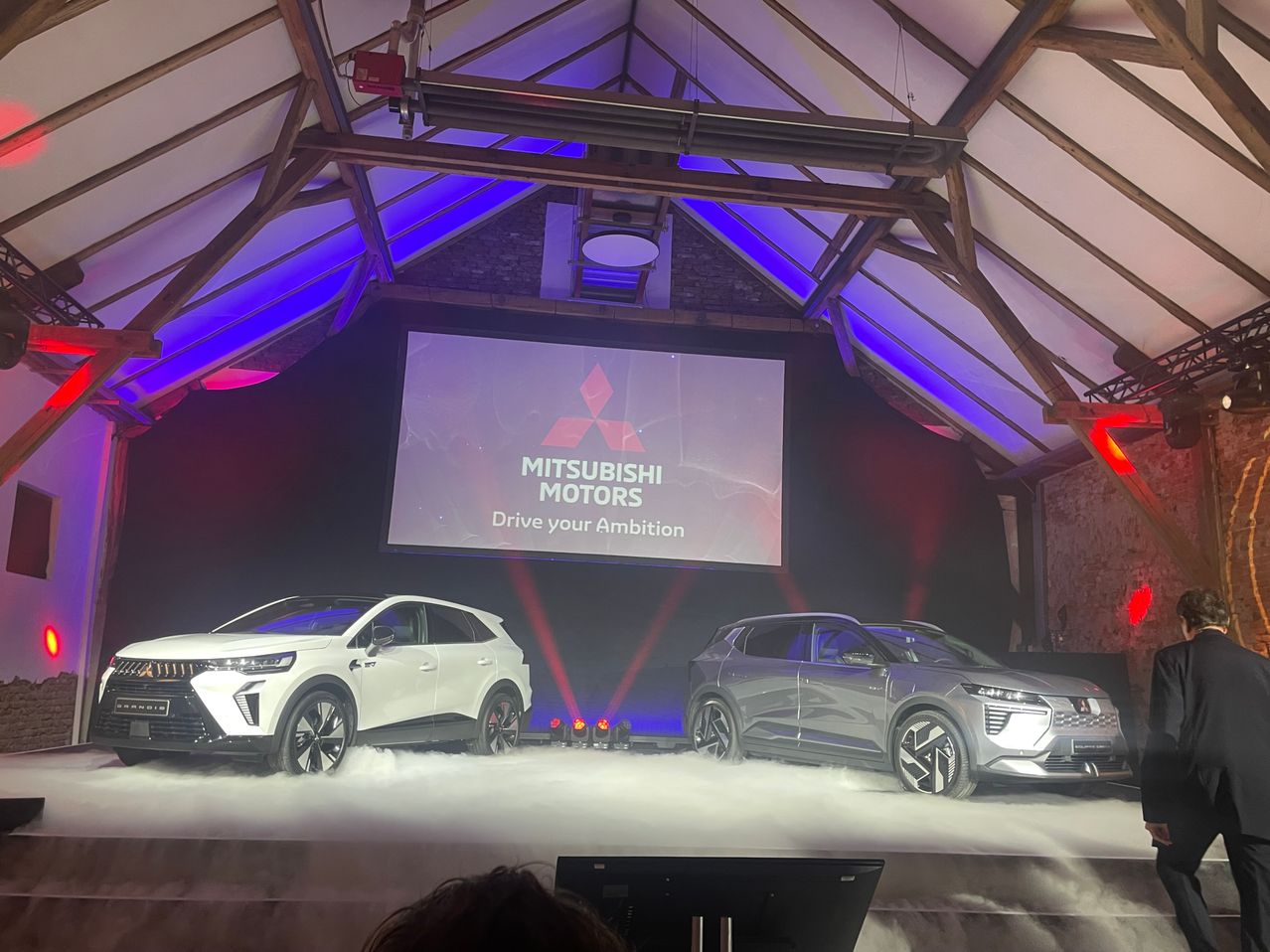 Zwei Mitsubishi SUVs stehen auf einer Bühne unter einem Mitsubishi Motors Banner mit dem Slogan Drive your Ambition.