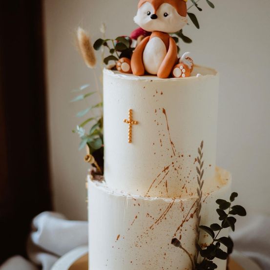 Ein dreistöckiger Kuchen mit weißer Glasur, braunen Spritzern und einem Fuchsaufsatz. Die oberste Etage hat ein goldenes Kreuz und grüne Blätter.