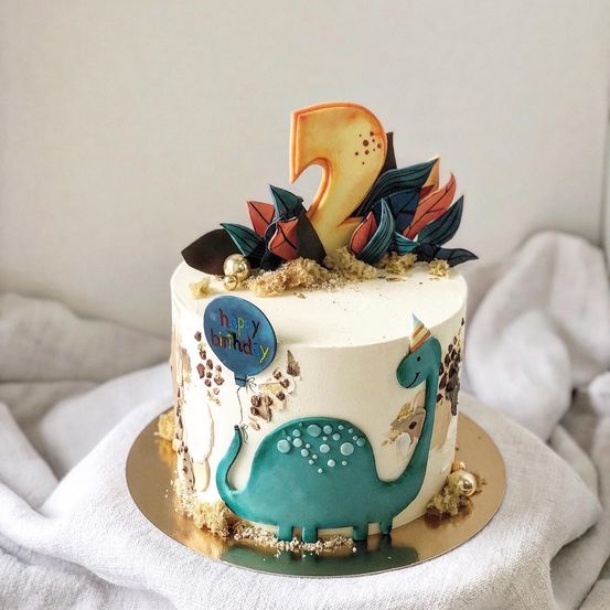 Ein weißer Kuchen mit einem grünen Dinosaurierdesign, einem Luftballon und einer Nummer 2 oben drauf. Er steht auf einem goldenen Teller.