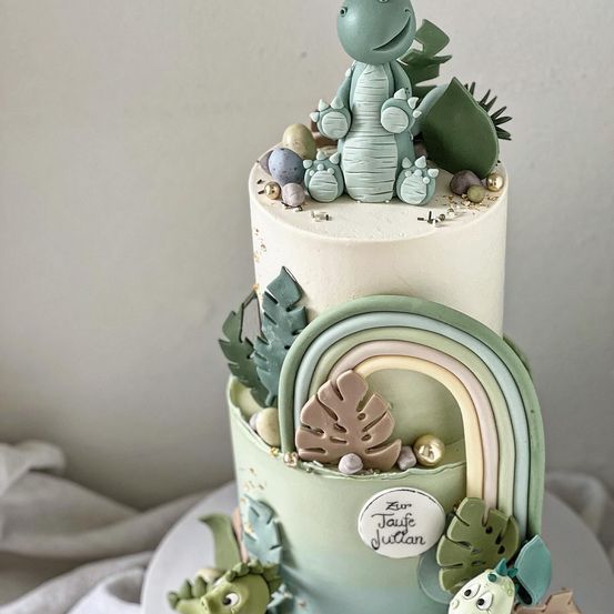 Ein zweistöckiger Kuchen, verziert mit einer Dinosaurierfigur, Blättern, einem Regenbogen und Eiern. Der Kuchen ist mit grüner Glasur verziert und hat ein Namensschild mit der Aufschrift 'Zur Taufe Julian.' Er steht auf einem weißen Teller.