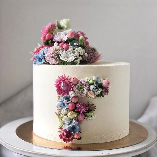 Ein weißer Kuchen mit einem Blumendesign in Form des Buchstabens 'F' auf einem goldenen Teller. Er hat Blumen darauf.