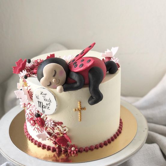 Ein Kuchen mit einer schlafenden Marienkäfer oben drauf, umgeben von roten Blättern und Blumen. Der Kuchen ist auf einem goldenen Teller mit einem weißen Tuch. Der Kuchen hat ein Schild mit den Worten 'Zur heiligen Taufe Marie.' Der Kuchen ist mit roten Perlen verziert.