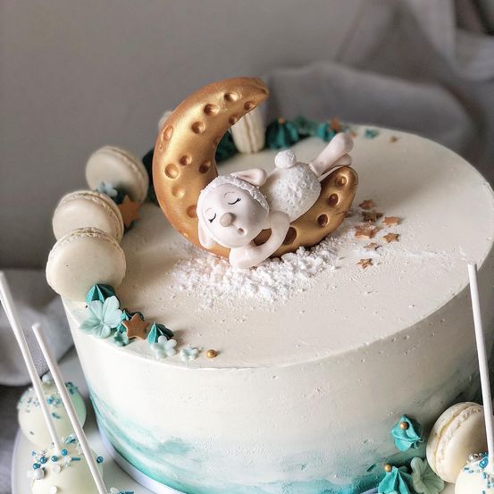 Ein Kuchen, der mit einem schlafenden Lamm, einem goldenen Halbmond und Streuseln aus weißen und blauen Blumen verziert ist. Macarons und Cake Pops sind um den Kuchen herum platziert.
