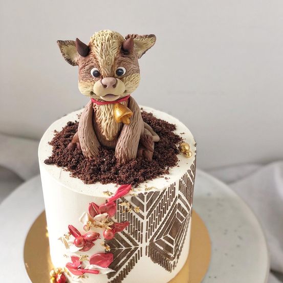 Ein Kuchen, verziert mit einem kleinen Tier, das in schokoladenüberzogener Erde sitzt. Der Kuchen hat eine weiße Zuckergussbasis mit braunen Mustern und roten und goldenen Dekorationen.