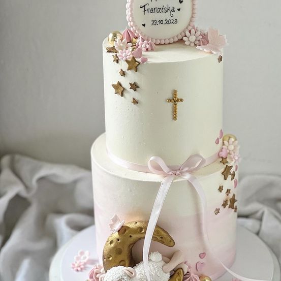 Ein dreistöckiger Kuchen mit rosa und weißer Thematik. Die oberste Schicht hat Sterne und ein Kreuz. Die mittlere Schicht hat ein rosa Band. Die unterste Schicht hat ein Lamm und einen Mond.